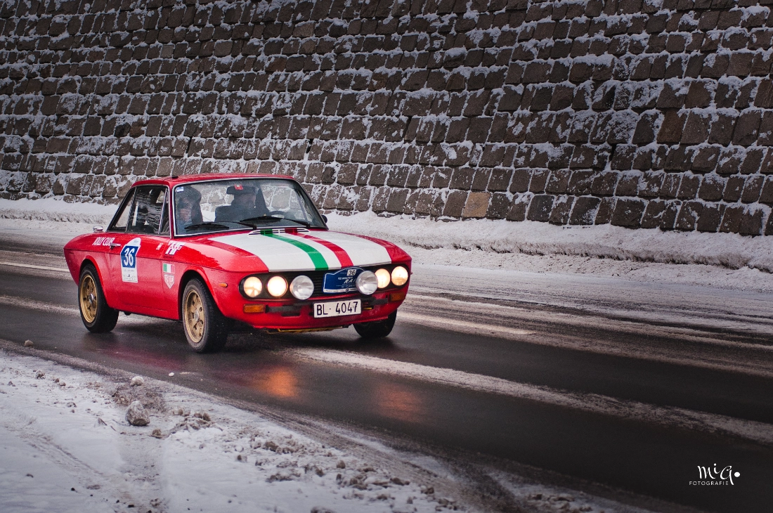 Ein roter klassischer Lancia Fulvia Rallyewagen fährt auf einer verschneiten Strasse an einer Steinmauer vorbei