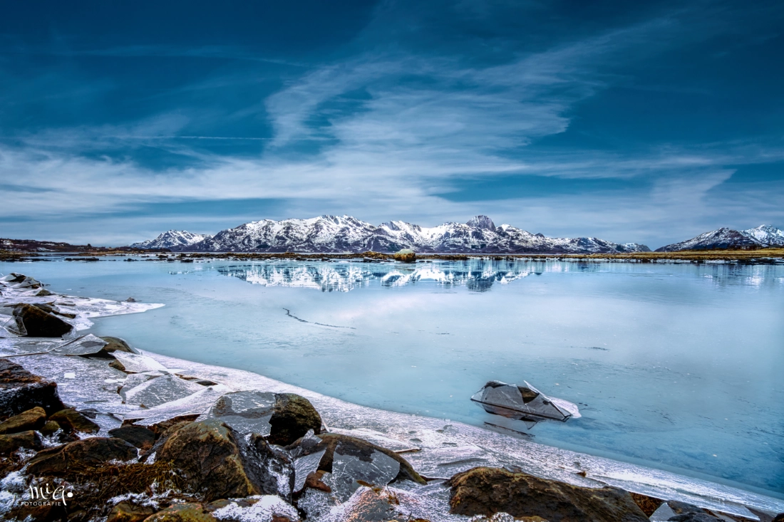 Teilweise zugefrorene Lagune auf Langøya in den Vesteraalen mit Eisschollenspiegelung der schneebedeckten Bergkette unter blauem Winterhimmel in Nordnorwegen