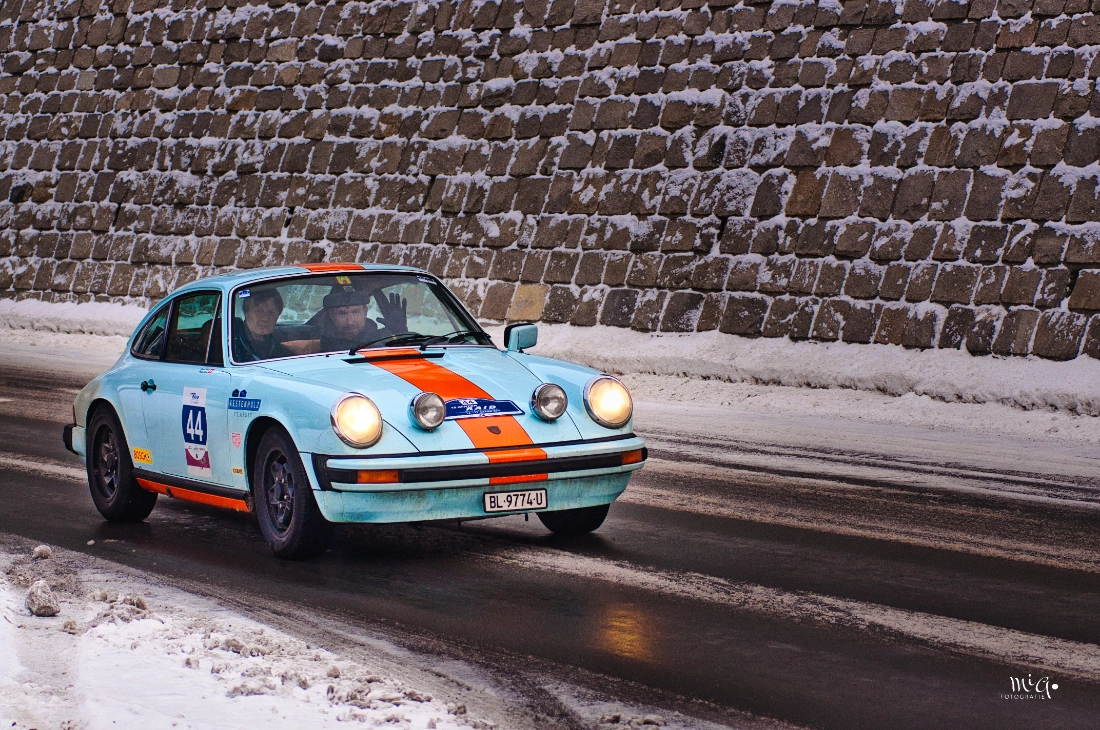 Ein hellblauer Porsche 911 mit orangen Streifen faehrt auf einer verschneiten Strasse an einer rustikalen Steinmauer vorbei