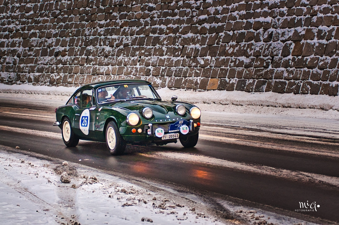 British Racing Green im Winterkleid Ein britischgrüner Triumph GT6 mit Startnummer 26 und Schweizer Kennzeichen fährt auf einer verschneiten Rallye-Strecke vor einer massiven Steinmauer