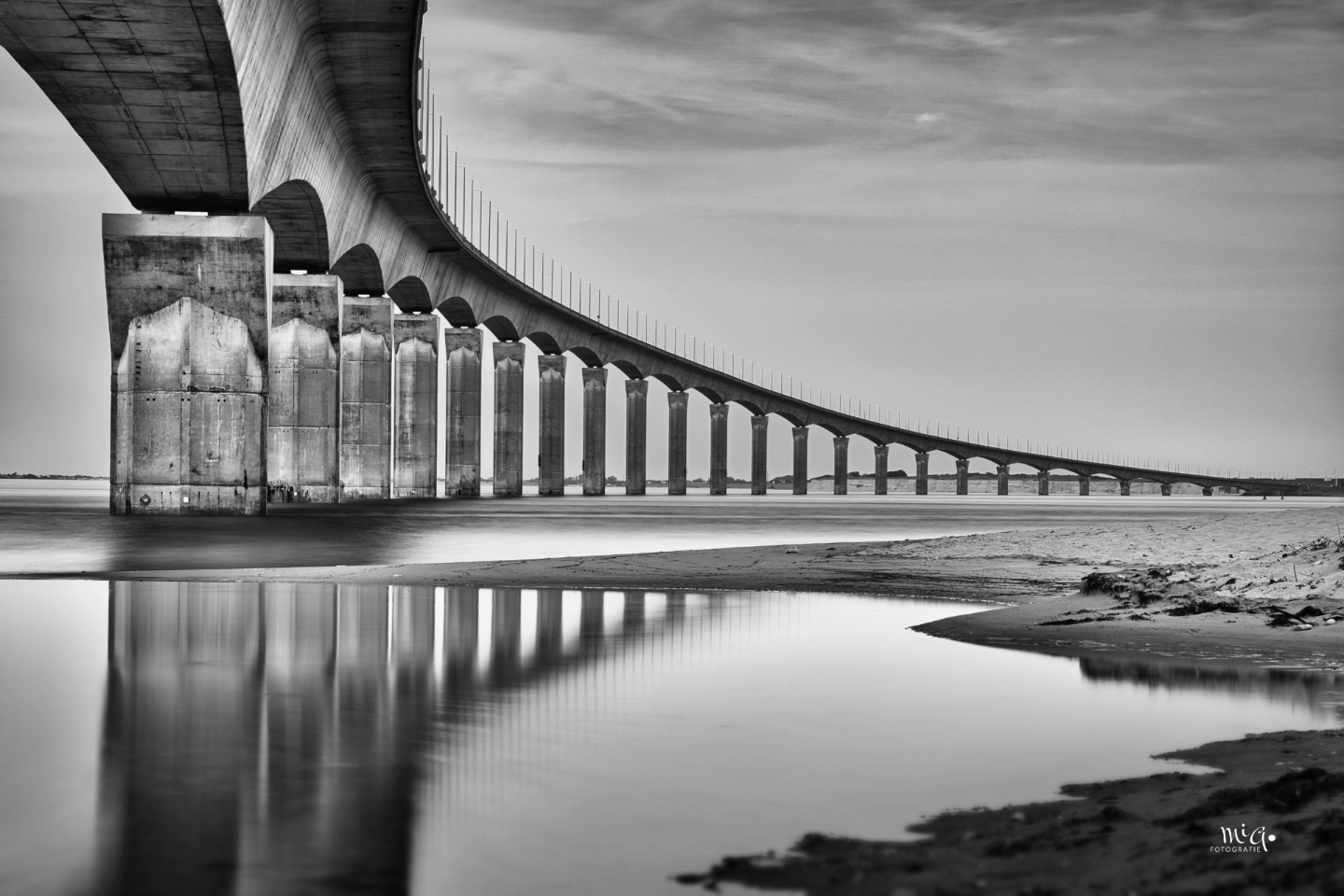 Schwarz-Weiss-Fotografie der Brücke zur Ile de Re (Pont de l'ile de Re) in Frankreich. Architekturaufnahme mit Fokus auf die Betonpfeiler, die sich im glatten Wasser spiegeln, unter einem leicht bewölkten Himmel.