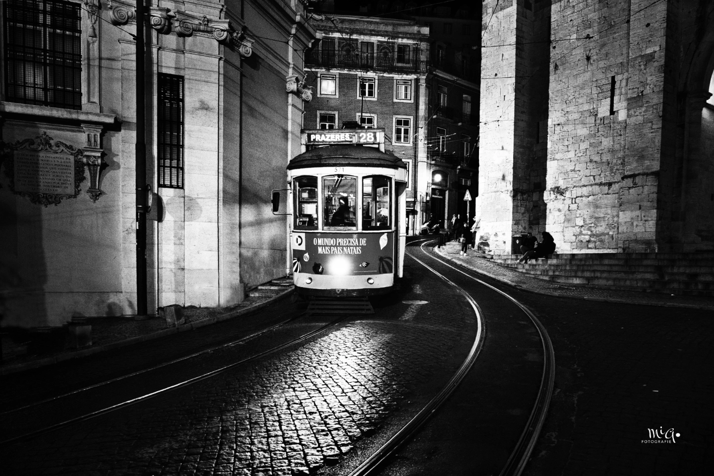 Schwarz-Weiss-Fotografie der historischen Strassenbahn Linie 28 in Lissabon bei Nacht. Die Bahn fährt auf Schienen durch eine enge Kurve zwischen historischen Gebäuden, beleuchtet von den eigenen Scheinwerfern auf Kopfsteinpflaster.