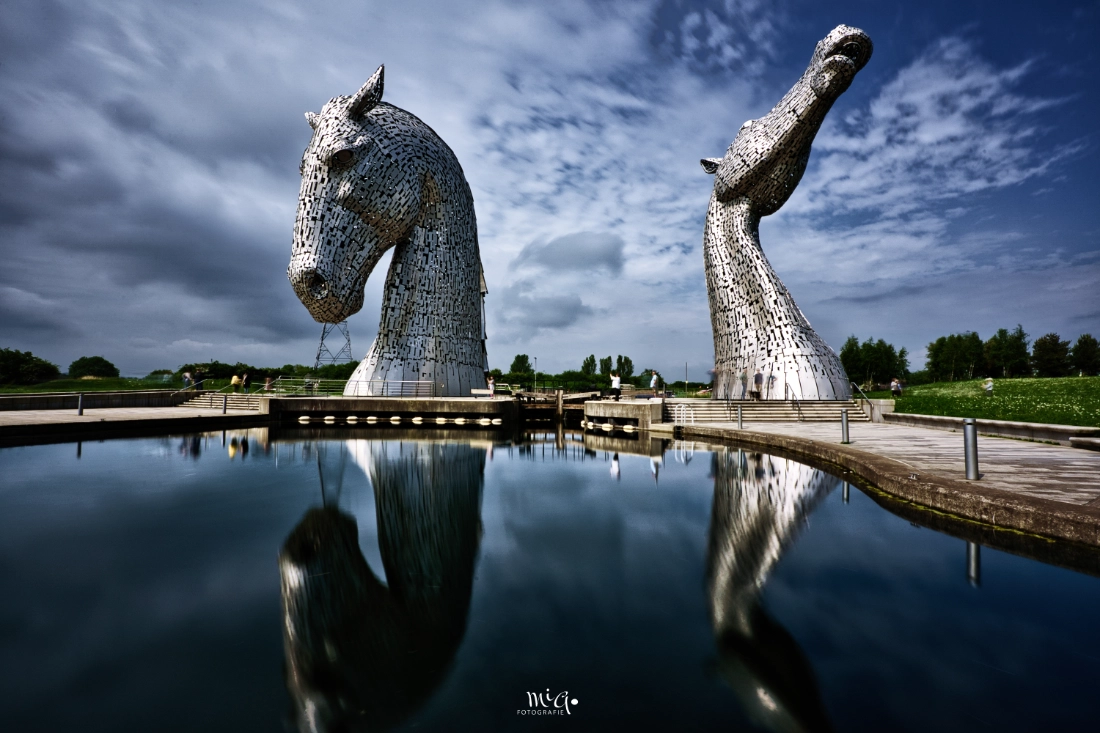 Die monumentalen Kelpies-Pferdekopfskulpturen aus Stahl in Falkirk, Schottland, unter einem wolkigen Himmel mit Spiegelung im Wasser.